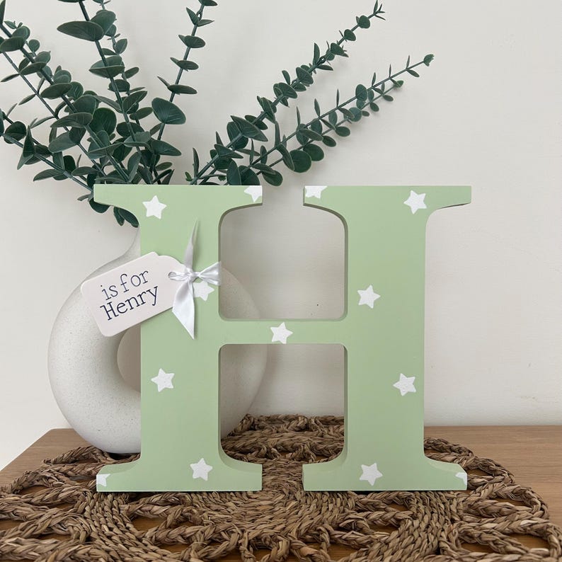 freestanding, bright green initial H with white stars on and a white tag attached to the left with a white ribbon with the words “is for Henry” on.