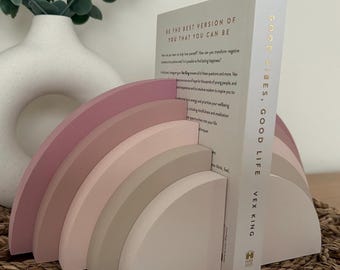 May include: A rainbow-shaped bookend set featuring five stacked semi-circular arches in graduated shades of dusty pink, mauve, and beige. A white book titled Good Vibes, Good Life by Vex King is positioned between the two halves of the bookend.
