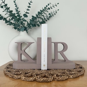 A pair of brown painted wooden bookends, shaped as the letters 'K' and 'R', supporting a book, with a white vase containing artificial greenery visible in the background, all on a woven surface
