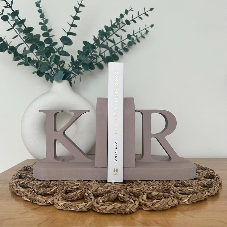 A pair of brown painted wooden bookends, shaped as the letters 'K' and 'R', supporting a book, with a white vase containing artificial greenery visible in the background, all on a woven surface