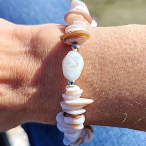May include: A bracelet made from natural shell and pearl beads. The design features a mix of shell and pearl elements, connected by silver-coloured metal beads. The shells are in shades of white, tan, and brown, creating a natural, organic aesthetic.