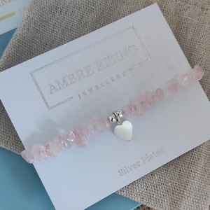 May include: A rose quartz bead bracelet with a silver heart charm. The bracelet is presented on a white card with the text "AMBRE RISING JEWELLERY" and "Silver Plated". The background includes a linen pouch and a blue envelope.