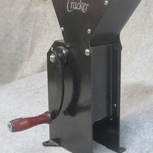 May include: Black metal "The Cracker" nutcracker with a hopper, hand crank, and a wooden handle. The nutcracker has a rectangular body with a flared top and is mounted on a base with two feet.