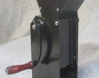 May include: Black metal "The Cracker" nutcracker with a hopper, hand crank, and a wooden handle. The nutcracker has a rectangular body with a flared top and is mounted on a base with two feet.