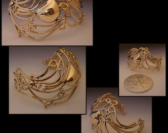May include: A gold-toned metal bracelet with a delicate, intricate design. The bracelet features a rounded, smooth center piece and a series of curved, flowing lines.