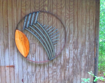 May include: A metal sculpture with a circular frame and a wooden oval shape in the center. The sculpture features a design of curved metal pieces radiating outward from the center.