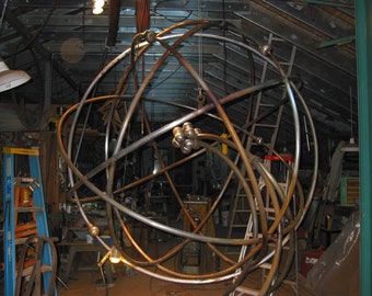 May include: A large metal sculpture of a sphere made of interconnected curved rods. The sculpture is suspended from the ceiling and appears to be in a workshop or studio setting.