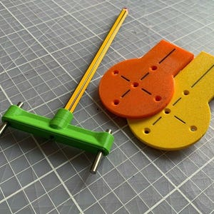 May include: A green plastic tool with a pencil inserted into it. The tool has two metal pins on either side. There are two circular orange and yellow plastic pieces with holes and lines on them.