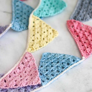 Puede incluir: Una guirnalda de banderines de crochet de colores vivos, con banderines en tonos azules, rosas, amarillos y verdes. Los banderines están conectados por una cuerda de crochet blanca.