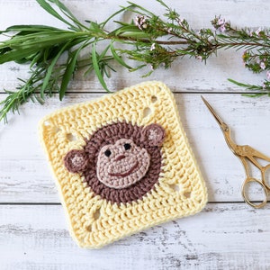 May include: A crocheted square with a brown monkey face in the center. The square is yellow and has a white background. There are green plants and a pair of gold scissors in the background.