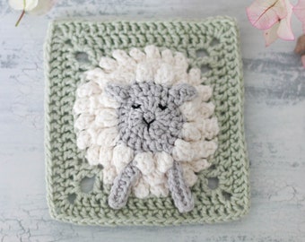 May include: A green crochet square with a white and gray crocheted sheep in the center. The sheep has a fluffy white body and a gray face with black eyes.