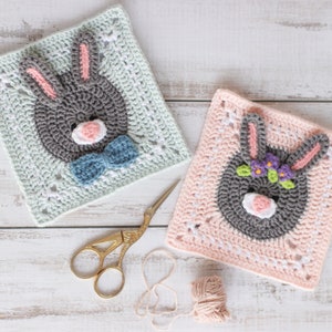 May include: Two crocheted bunny squares, one in light blue with a gray bunny wearing a blue bow tie, and the other in pink with a gray bunny wearing a flower crown.  The squares are on a white wooden surface with a pair of gold scissors and a ball of pink yarn.