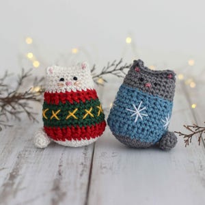 May include: Two crocheted cat ornaments wearing festive sweaters. One cat is white with a red and green sweater with a snowflake pattern. The other cat is gray with a blue sweater with a snowflake pattern.