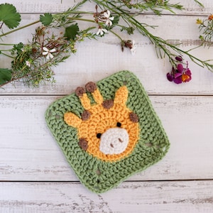 May include: A crocheted square featuring a giraffe face in yellow, brown, and white yarn. The square is surrounded by green yarn and is lying on a white wooden surface.