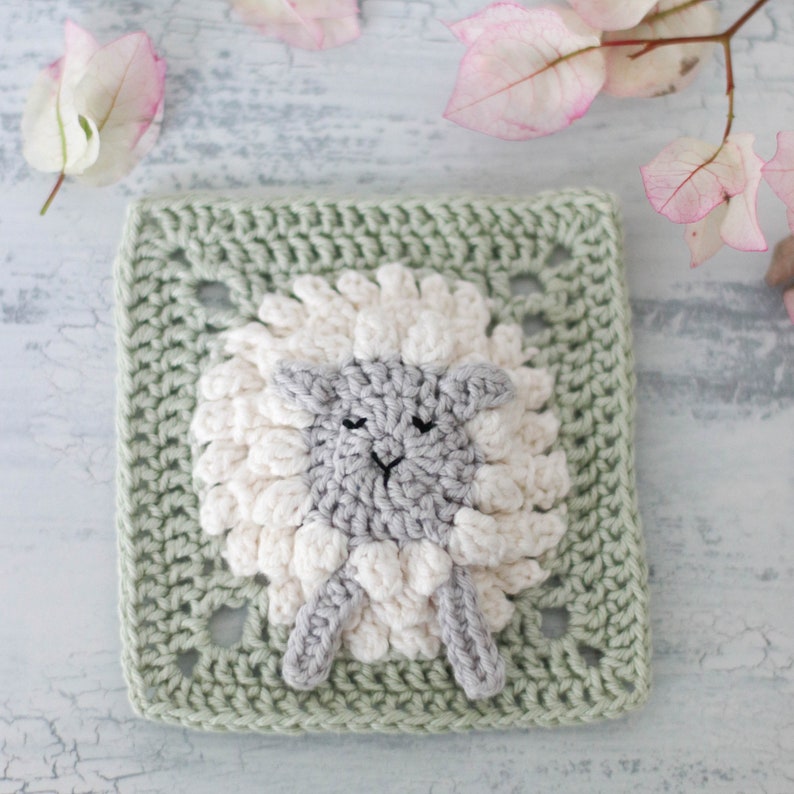 A 3D crochet granny square with a sheep / lamb motif. The square is mint green and the crochet lamb is white and grey. The square is surrounded by pink flowers.