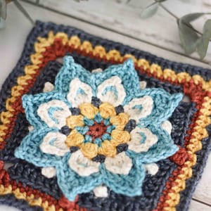 Puede incluir: Un cuadrado tejido a crochet con un diseño de flor azul, blanco, amarillo y rojo. La flor está rodeada por un borde azul oscuro con acentos amarillos, naranjas y rojos.
