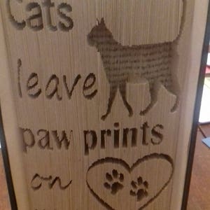 May include: A book with the words "Cats leave paw prints on your" carved into the pages. The pages are folded to create a silhouette of a cat and a heart with paw prints.