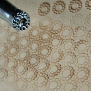 May include: Close-up of a leather stamping tool with a circular design. The tool is being used to create a pattern of small circles on a piece of leather.