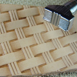 May include: A close-up image shows a leather craft tool creating a basketweave pattern on light tan leather. The metal stamp is in the upper right, revealing its detailed texture. The leather has a textured surface, with the pattern clearly defined.
