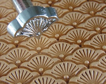 May include: A close-up of a metal leather stamping tool with a fan-shaped design. The tool is being used to create a pattern on a piece of leather.