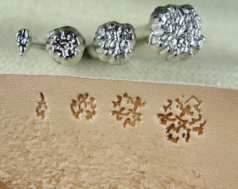 May include: Four metal stamps with intricate designs, used for leather crafting. The stamps are arranged in a row from left to right, with the largest stamp on the right. The stamps are shown on a piece of leather with impressions of the stamps.