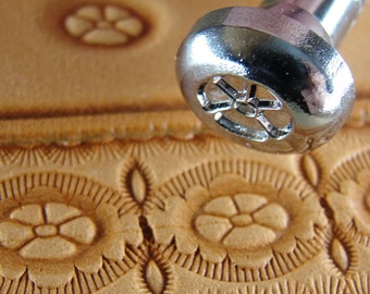 May include: Close-up of a silver leather stamping tool with a flower design. The tool is being used to create a pattern on a piece of light brown leather.