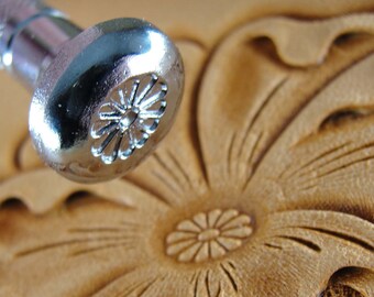 May include: Close-up of a silver leather stamping tool with a flower design. The tool is being used to create a flower design on a piece of leather.
