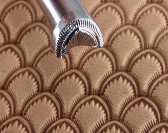 May include: Close-up of a silver leather stamping tool with a scalloped design. The tool is resting on a piece of brown leather that has been embossed with the same design.