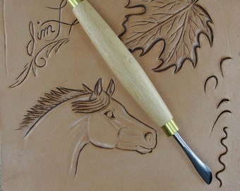 May include: A leather carving tool with a wooden handle and a curved metal blade. The tool is lying on a piece of leather that has been carved with a horse, a leaf, and a swirl design. The leather is light brown in colour.