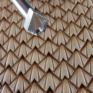 Pode incluir: Um close-up de uma ferramenta de carimbo de couro com um padrão decorativo de triângulos sobrepostos. A ferramenta é feita de metal e tem uma alça de madeira. A ferramenta é usada para criar um padrão em um pedaço de couro.