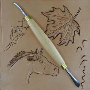 May include: A leather carving tool with a wooden handle and a curved metal blade. The tool is lying on a piece of leather that has been carved with a horse, a leaf, and a swirl design. The leather is light brown in color.