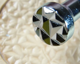 May include: Close-up of a silver-toned metal knob with a geometric, star-shaped design. The knob is in focus, while the background is blurred.