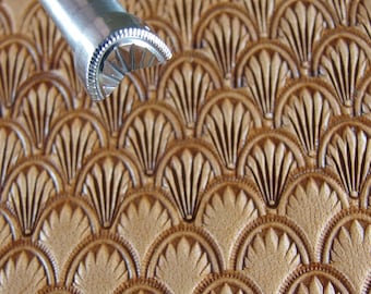 Op de afbeelding: Close-up van een metalen leerstempelgereedschap naast een stuk gestempeld leer. Het leer is bruin en heeft een herhalend waaiervormig patroon. De tool is zilverkleurig en heeft een gebogen, patroon stempeloppervlak.