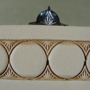 Puede incluir: Un pin decorativo plateado con un dise&ntilde;o geom&eacute;trico, sobre una superficie beige claro texturizada. Debajo, un patr&oacute;n repetitivo de c&iacute;rculos y motivos florales en un beige m&aacute;s oscuro a&ntilde;ade detalles.