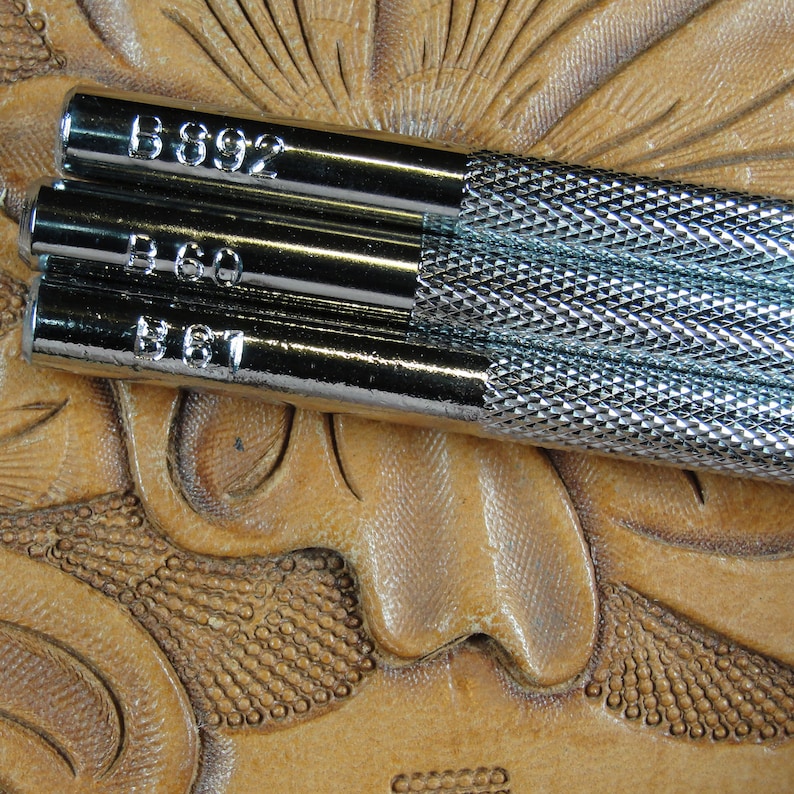 Puede incluir: Primer plano de tres herramientas met&aacute;licas para trabajar el cuero con mangos texturizados y n&uacute;meros estampados: B892, B60 y B61. Las herramientas descansan sobre una superficie de cuero marr&oacute;n claro tallada con dise&ntilde;os florales. Las herramientas son plateadas y brillantes.