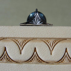 Puede incluir: Un objeto decorativo de metal plateado con un dise&ntilde;o geom&eacute;trico, colocado sobre una superficie texturizada de color blanco roto. El objeto tiene forma triangular con un elemento central puntiagudo y una serie de l&iacute;neas radiales. Debajo, un patr&oacute;n repetitivo de arcos estilizados.