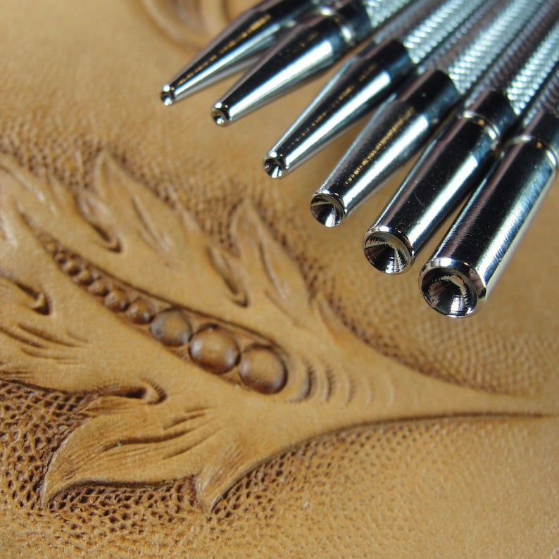 Peut inclure: Un ensemble de six outils de travail du cuir argent&eacute;s avec des pointes de diff&eacute;rentes tailles. Les outils sont dispos&eacute;s en &eacute;ventail et reposent sur un morceau de cuir brun avec un motif d&eacute;coratif.