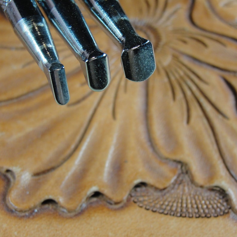 Puede incluir: Primer plano de tres herramientas de marroquiner&iacute;a met&aacute;licas con cabezas de diferentes formas. Las herramientas est&aacute;n colocadas sobre una superficie de cuero marr&oacute;n claro con un dise&ntilde;o floral. El cuero ha sido trabajado con l&iacute;neas y texturas detalladas.