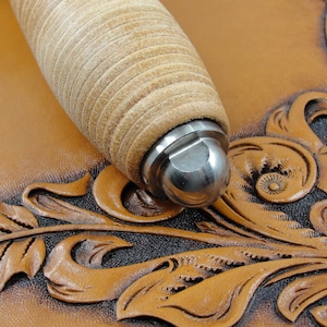 May include: Close-up of a leather tool with a wooden handle and a metal tip. The tool is resting on a piece of brown leather with an intricate floral design.