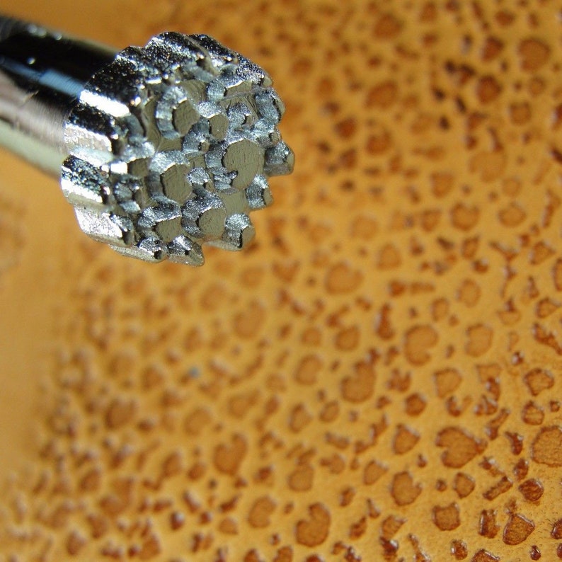Op de afbeelding: Close-up van een zilveren metalen stempel met een gedetailleerd, gestructureerd ontwerp. De stempel wordt gebruikt voor het bewerken van leer.