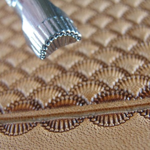 May include: Close-up of a metal leather stamping tool with a scalloped edge. The tool is being used to create a decorative pattern on a piece of brown leather.