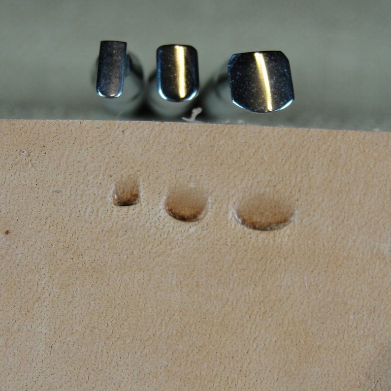 Puede incluir: Tres herramientas met&aacute;licas para estampar cuero con cabezas de diferentes formas se muestran sobre tres impresiones en un trozo de cuero marr&oacute;n claro. Las herramientas son brillantes y reflejan la luz. Las impresiones est&aacute;n espaciadas uniformemente y muestran el efecto de las herramientas.