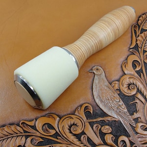 May include: A white leather mallet with a wood handle, used for leather crafting. The mallet is resting on a brown leather surface with an embossed design of a bird and floral patterns.