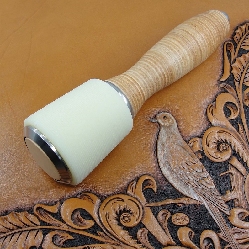 May include: A white leather mallet with a wood handle, used for leather crafting. The mallet is resting on a brown leather surface with an embossed design of a bird and floral patterns.