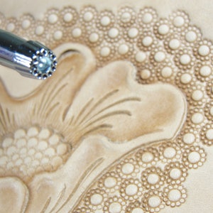 May include: A close-up of a leather stamping tool with a circular design. The tool is being used to create a floral pattern on a piece of leather.