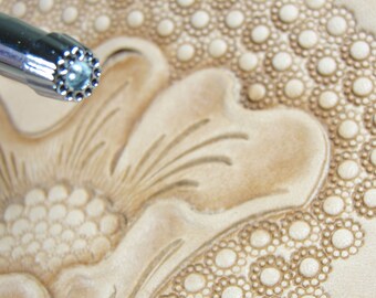 May include: A close-up of a leather stamping tool with a circular design. The tool is being used to create a floral pattern on a piece of leather.