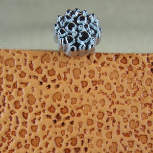 Op de afbeelding: Een decoratieve stempel van zilverkleurig metaal met een bloemmotief. De stempel ligt op een bruin lederen oppervlak.