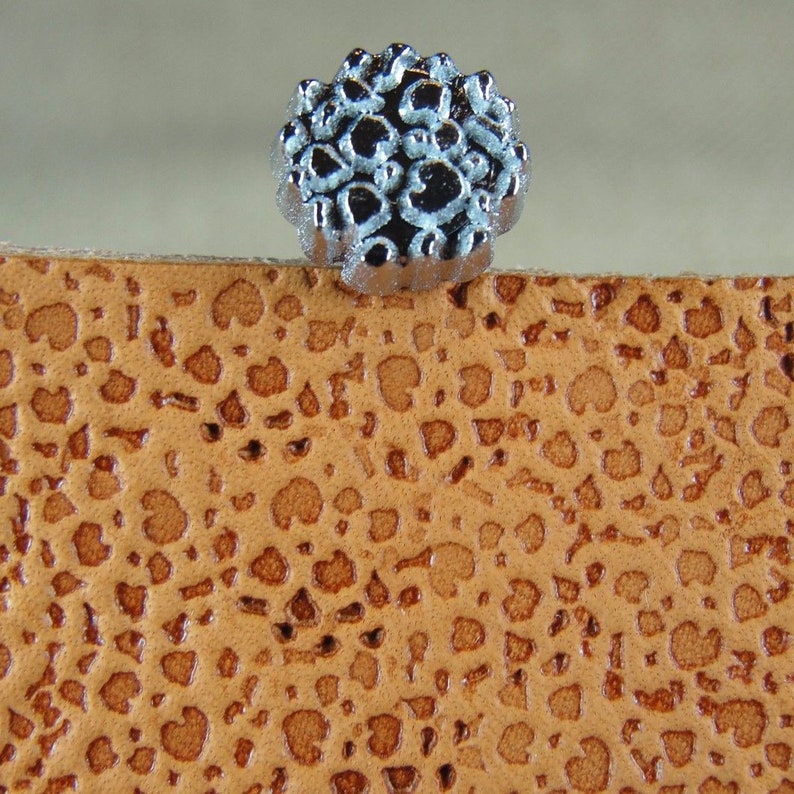Op de afbeelding: Een decoratieve stempel van zilverkleurig metaal met een bloemmotief. De stempel ligt op een bruin lederen oppervlak.