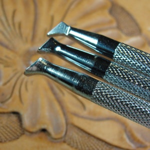 Puede incluir: Tres herramientas plateadas para trabajar el cuero, con mangos texturizados y cuchillas anguladas, dispuestas en diagonal. Las herramientas se colocan sobre un fondo borroso de cuero tallado, sugiriendo un tema artesanal o de afici&oacute;n.