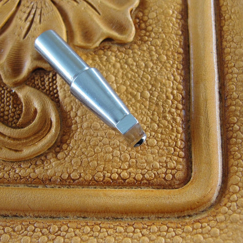 Puede incluir: Una herramienta de trabajo del cuero de metal plateado con una punta puntiaguda, que descansa sobre una superficie de cuero marr&oacute;n con un patr&oacute;n texturizado.